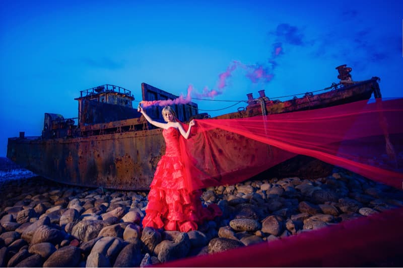 這張照片是一個穿著紅色禮服的女性站在一艘生鏽的船旁邊，她拿著一支燃燒的紅色煙霧彈，在藍色的夜空下，鮮紅色的煙霧與她的禮服交相輝映，形成一道美麗的風景線。