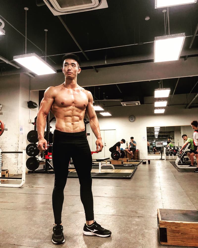 這張「健身房」的照片是一個健美男性站在健身房裡，他穿著黑色的緊身褲和運動鞋，並展示著他強壯的肌肉。照片的背景是健身房的器材，包括槓鈴、啞鈴和鏡子。