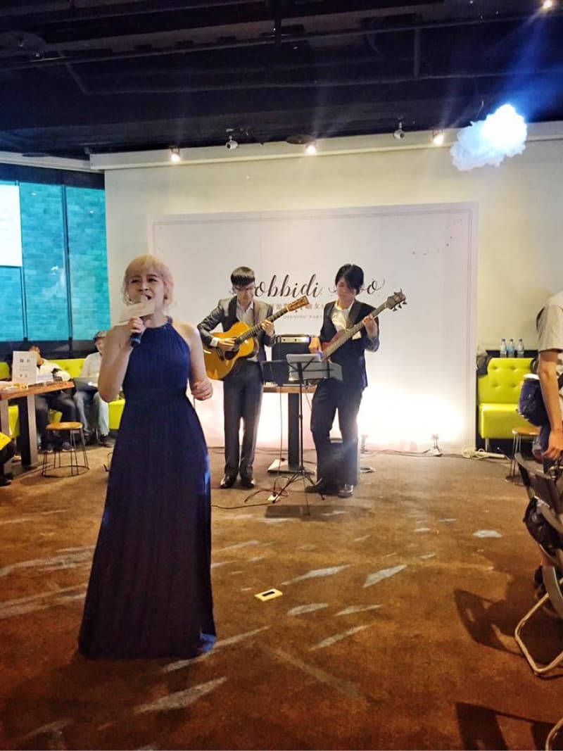 這張照片是一個小型音樂表演，一位穿著藍色禮服的女性歌手正在唱歌，她的身後有兩位男性樂手，一位彈吉他，另一位彈貝斯。