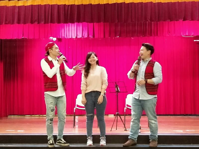 這張「舞臺表演」的照片是三位主持人站在舞臺上,一位女性主持人穿著淺色上衣和牛仔褲,兩位男性主持人穿著紅色背心,他們手上都拿著麥克風,正在進行舞臺表演。