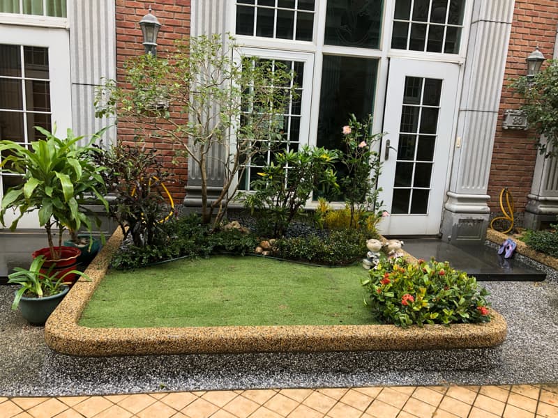 這張照片是一個花園，花園中間有一個用小石頭圍成的方形草坪，草坪周圍種滿了綠色植物和花草。