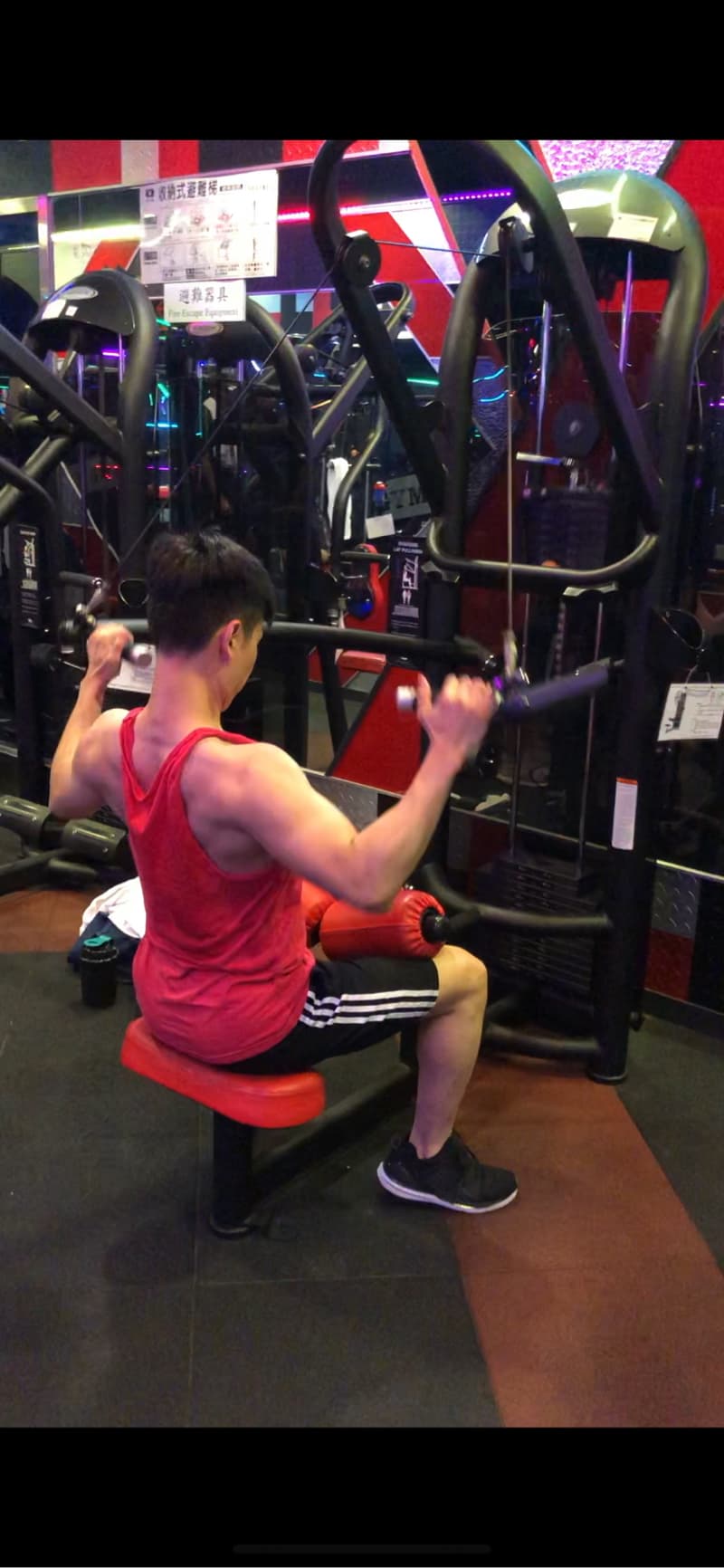 這張照片是一名男子在健身房使用拉力器鍛鍊背部肌肉。他正坐在拉力器上，雙手握住拉力器的手柄，並向後拉動，使背部肌肉得到鍛鍊。他穿著紅色的背心和黑色的運動短褲，戴著一頂黑色的帽子，看起來非常專注。