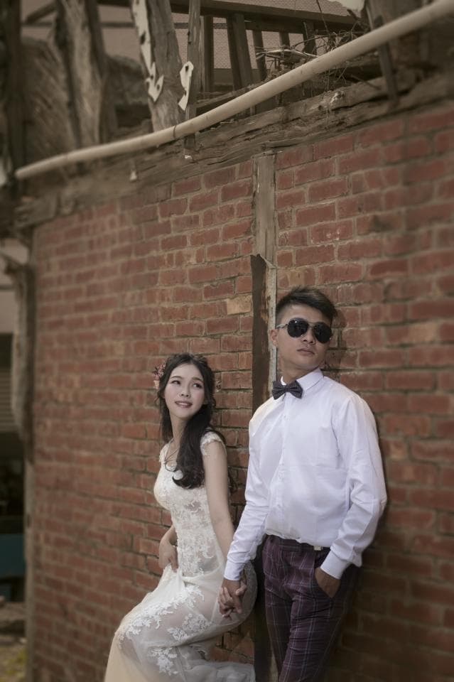 這張照片是 一對情侶在紅磚牆前合照。女方穿著白色蕾絲婚紗,男方穿著白色襯衫和紫色西裝褲,兩人笑著看著鏡頭,畫面溫馨浪漫。