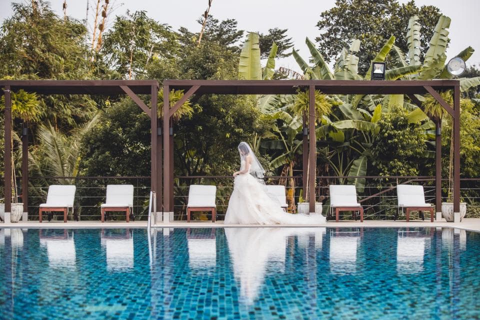 這張「婚禮」的照片是一個穿著婚紗的新娘,在一個有陽光和棕櫚樹的泳池邊走著,她正在走向一個木質涼亭。