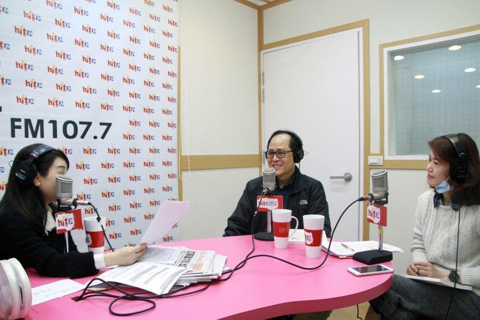 這張照片是三個人在一個廣播室裡,他們坐在一張粉紅色的桌子前,面前放著麥克風,正在錄製節目。