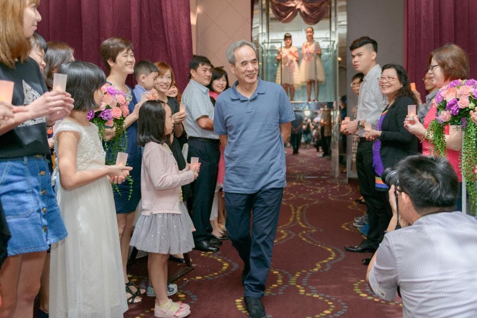 這張婚禮照片是一個婚禮場景,裡面有許多參加婚禮的賓客,他們穿著得體的服裝,並且在婚禮現場拍照。其中一位男性賓客站在人群中,顯得特別突出。他穿著藍色襯衫,手裡拿著一個酒杯,看起來非常開心。