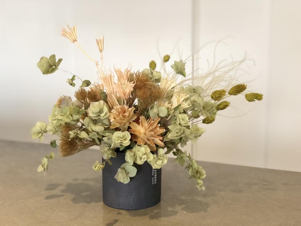 1) 照片顯示一個灰色花瓶,裡面裝滿了乾燥花,包括綠色和棕色的花朵以及一些白色的花朵,花瓶放在一個木質的檯面上,背景是白色的牆壁