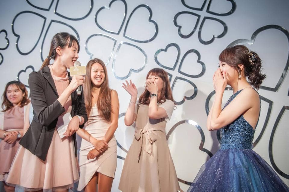 這張「婚禮」的照片是一群朋友在慶祝婚禮,他們站在白色牆壁前,背景是四葉草的裝飾。他們都很開心,其中一個女孩還拿著麥克風,看起來像是正在發表感言。