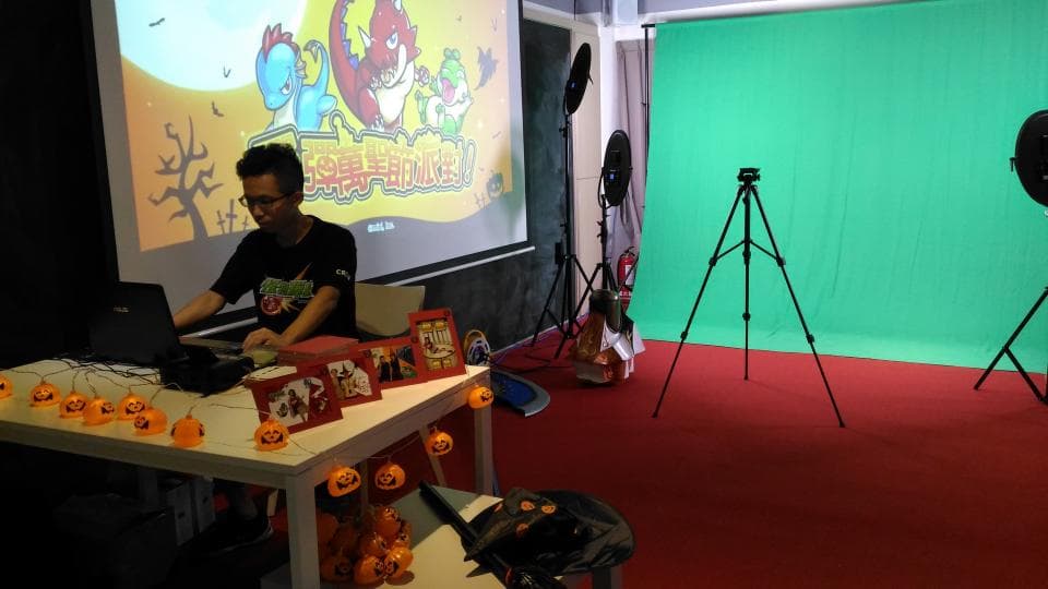 這張「影片拍攝」的照片是一名男子在綠幕前使用電腦工作,地上有腳架和燈光設備。