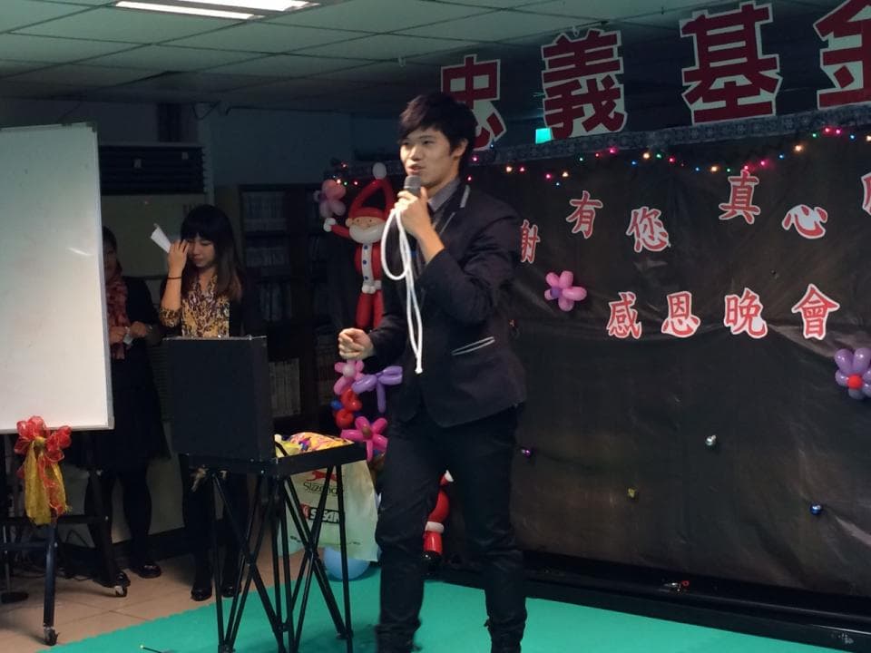 這張照片是一個男人在表演魔術。他站在一個黑色小桌旁,手上拿著一個白色繩子,看起來像是在表演一個神奇的魔術把戲。