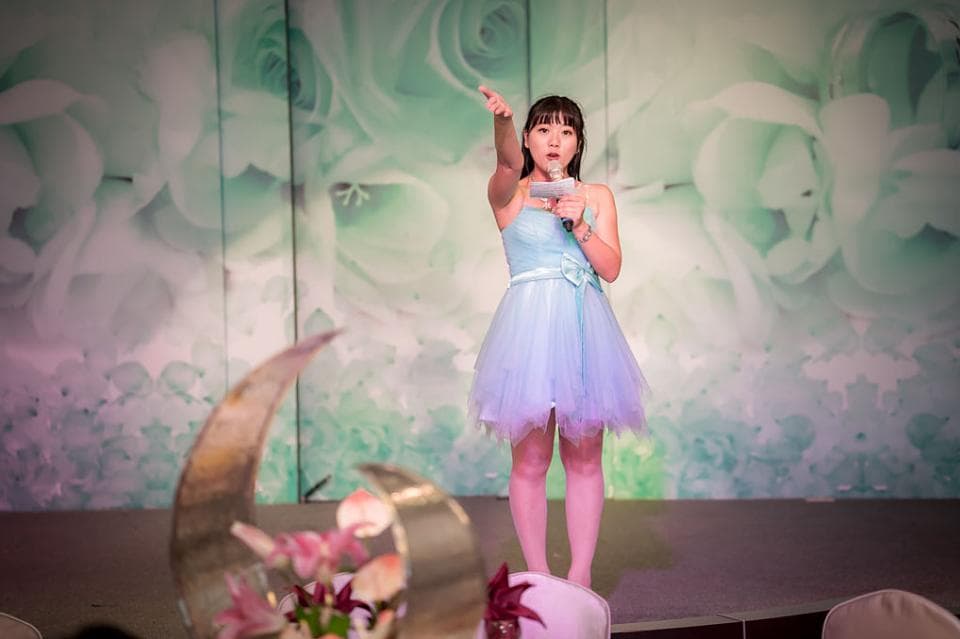 這張「婚禮」的照片是一名穿著淺藍色禮服的女性,站在舞臺上拿著麥克風說話,背景是綠色的花卉圖案。