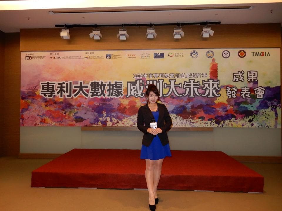 這張「展覽宣傳照片」是一個年輕女子站在一個有舞臺的房間裡,她穿著藍色的連衣裙,站在一個紅色的舞臺上,背景是一個大型的展覽宣傳海報。