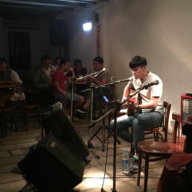 這張照片是一個音樂表演,最重要的是唱歌的男生,他坐在椅子上,拿著吉他,正專注地唱歌。