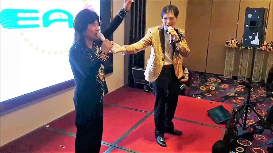 這張「兩位男士在舞臺上唱歌」的照片是,一位穿著黑色西裝外套的男士正在唱歌,另一位穿著金色西裝外套的男士拿著麥克風在旁邊。舞臺上有一臺音響和一個麥克風架。