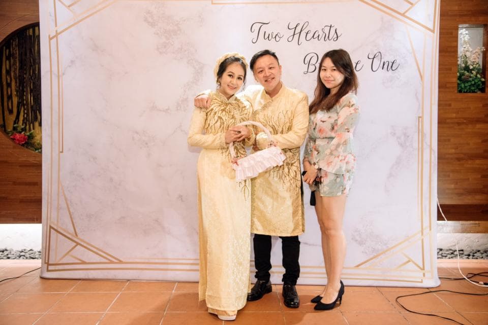 這張照片是三個人在婚禮背景板前合影,中間是新郎,穿著金色的西裝,左邊是新娘,穿著米白色的婚紗,右邊是伴娘,穿著粉色的禮服。