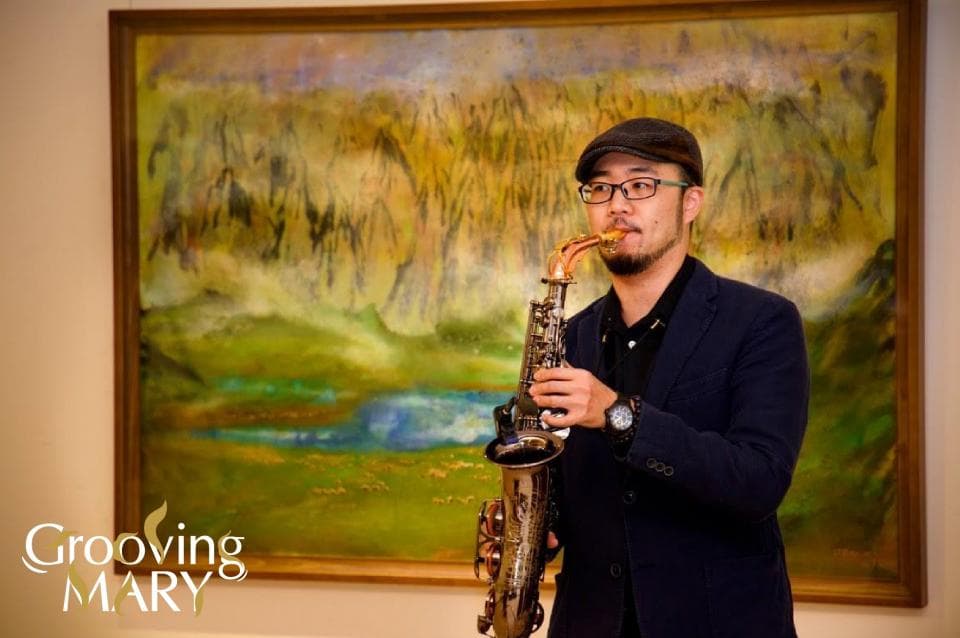 這張照片是一個男人站在一幅風景畫前,他拿著一把薩克斯風,似乎準備演奏。