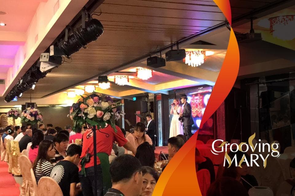 這張婚宴的照片是一個婚禮現場,有新郎新娘在臺上,臺下坐著許多賓客。舞臺上還有燈光和音響設備,賓客們正在享用美食和飲品。