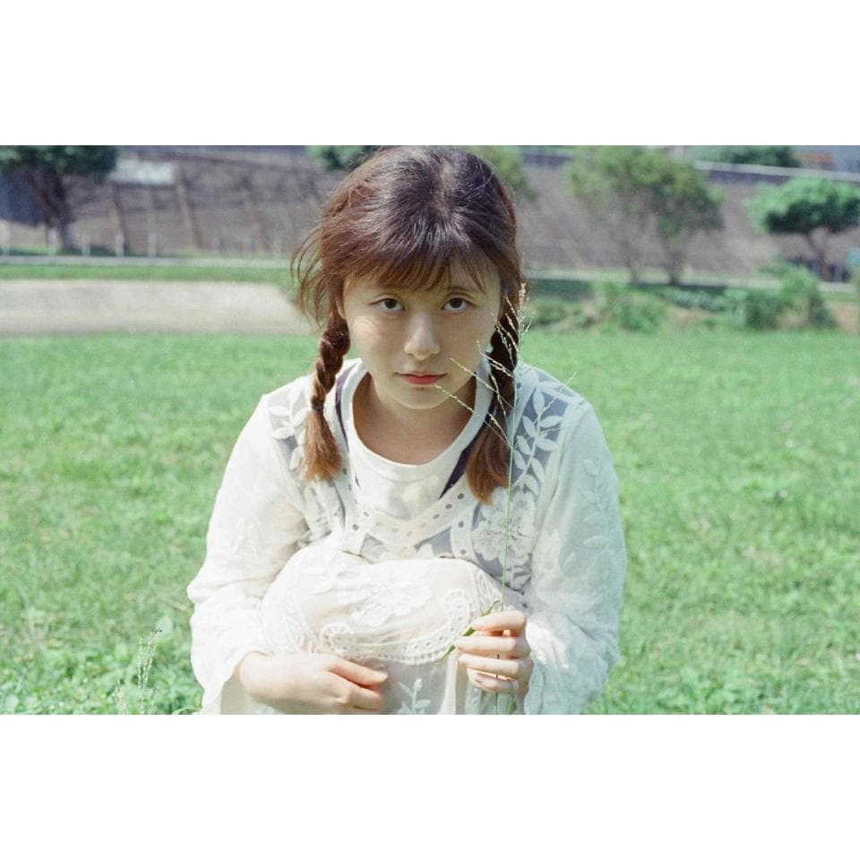 這張照片是一個年輕女孩在草地上穿著白色蕾絲衣服的肖像。她有著可愛的臉龐和一頭烏黑亮麗的長髮,正用溫柔的眼神看著鏡頭,散發著青春的氣息。