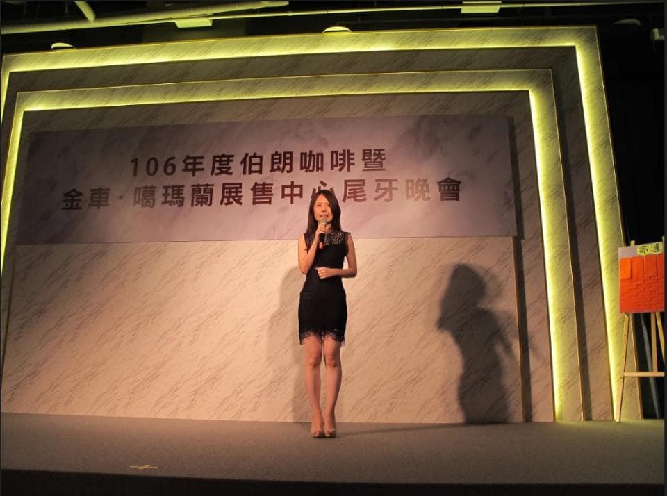 這張照片是一個舞臺,舞臺上有一位女士在演講,她穿著黑色的洋裝,拿著麥克風,正對著觀眾說話。