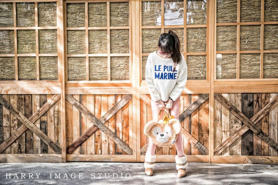 這張「女生站在木頭牆前」的照片是,女生穿著白色上衣,背著米色毛絨玩具,站在木頭牆前,牆面由許多格柵組成。