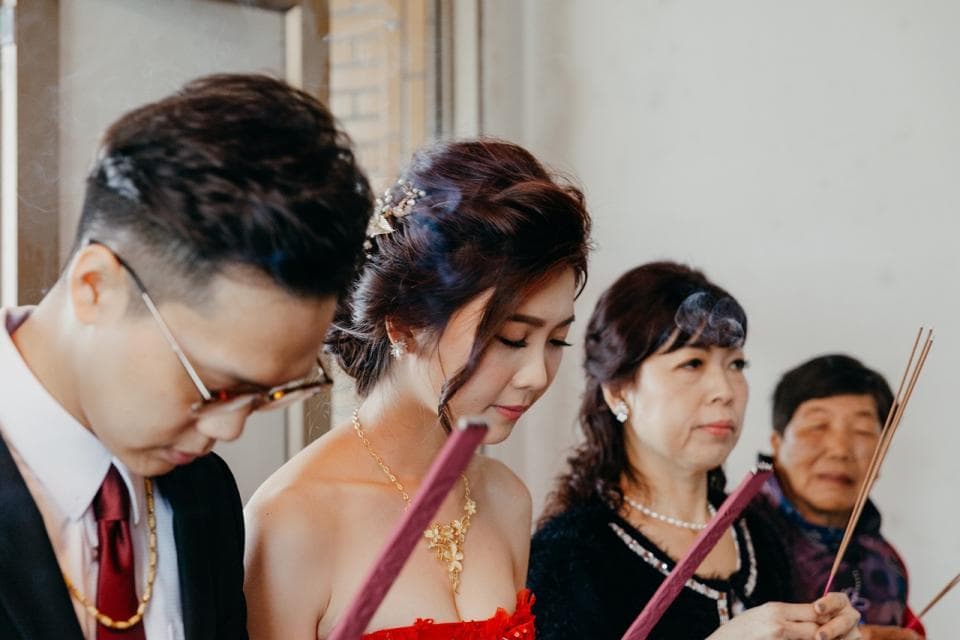 這張照片是新郎新娘在婚禮儀式上燒香。新娘身穿紅色禮服,頭戴花環,笑容甜美,新郎則站在她身旁,表情溫柔地看著她。他們面前的香爐冒著白煙,象徵著美好的祝福。