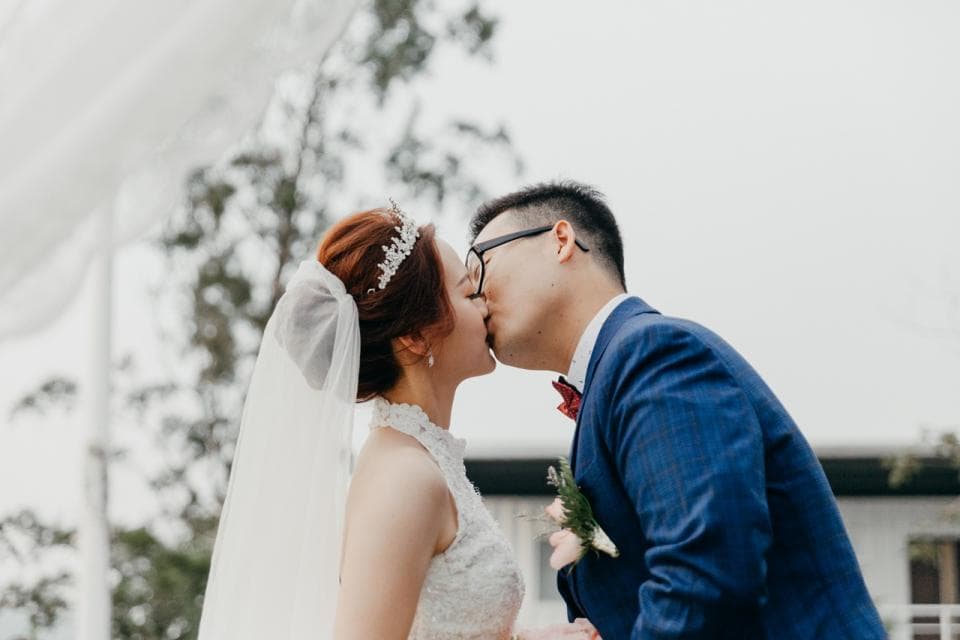 這張照片是一對新人正在接吻。新郎穿著深藍色的西裝,戴著眼鏡,新娘穿著白色婚紗,戴著皇冠。他們看起來非常幸福。