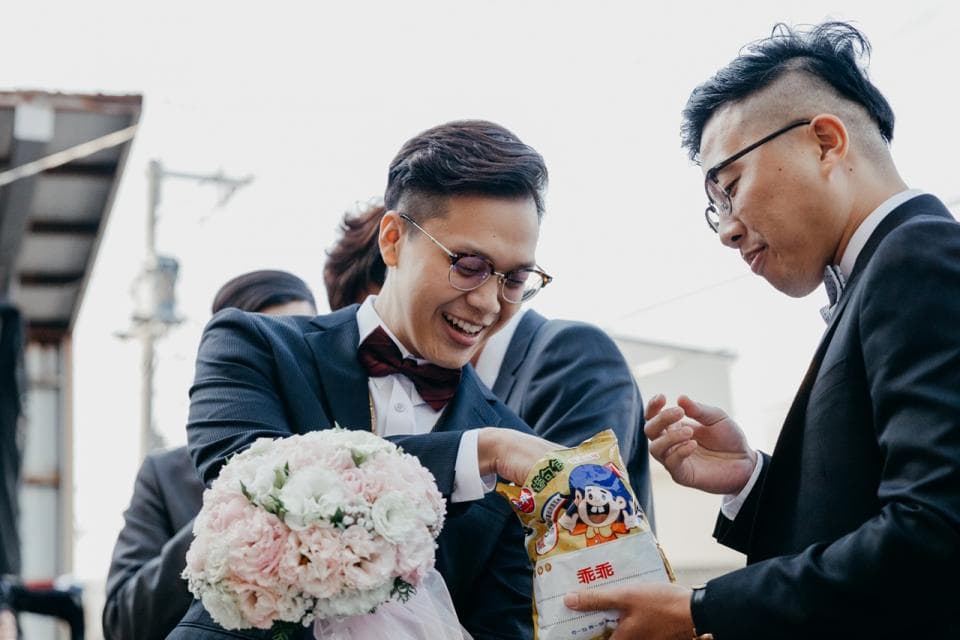 這張照片是一個新郎在結婚儀式上接受祝福。他穿著西裝,打著領結,手裡拿著一束花,笑容滿面。他旁邊是一位親友,正要給他一個紅包。
