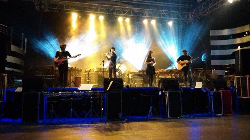 這張照片是一個音樂表演的舞臺,舞臺上有四個音樂家,兩個彈吉他,一個吹長笛,一個唱歌。他們站在一個亮麗的舞臺上,背後有強烈的燈光,讓他們更顯眼。