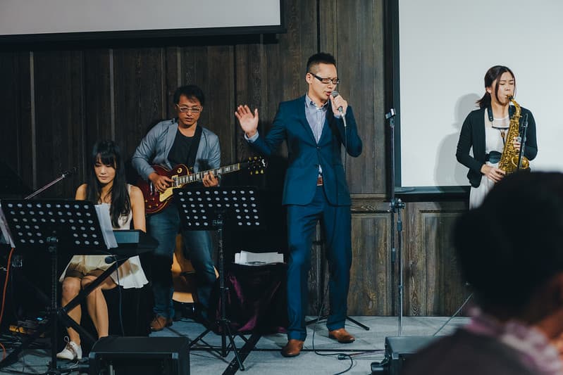 這張「婚禮樂隊」的照片是,一個穿著西裝的男人正在唱歌,另一個男人正在彈吉他,還有一位女人在演奏薩克斯風,以及一位女人在彈奏鍵盤。