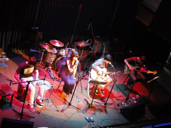 這張照片是四個年輕人在舞臺上演奏音樂。 左邊的男生戴著帽子,正在敲打鼓。 右邊的女生穿著紅色的衣服,正在唱歌,她的手拿著麥克風。 後面的兩個男生正在彈吉他,他們看起來很享受音樂。