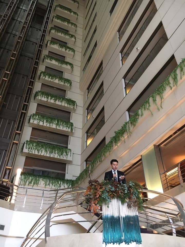 這張「酒店大廳」的照片是一名男子在一個裝飾著綠色植物和藍色流蘇的平臺上,手持麥克風,似乎正在發表演講。