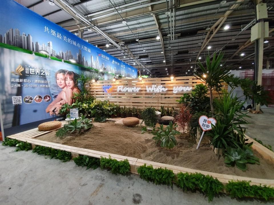 這張「展覽攤位」的照片是,一個以木板搭建的沙灘景觀,配有綠色植物、仿真沙子以及木質座椅,牆壁上有廣告牌和霓虹燈裝飾。