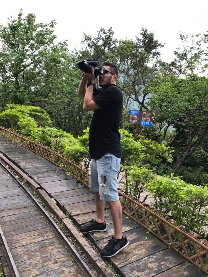 這張「男人在山路旁拍照」的照片是,一位男人站在一條小路旁,他拿著相機在拍攝風景。他穿著牛仔短褲和運動鞋,看起來很休閒。