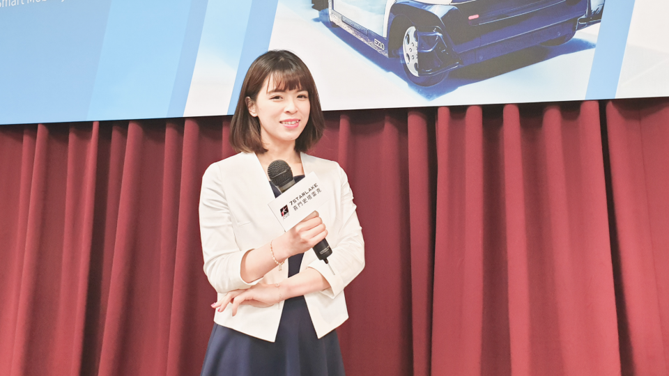 這張照片是一名女性主持人站在舞臺上,她穿著白色外套和藍色連身裙,手上拿著麥克風,背景是紅色窗簾。