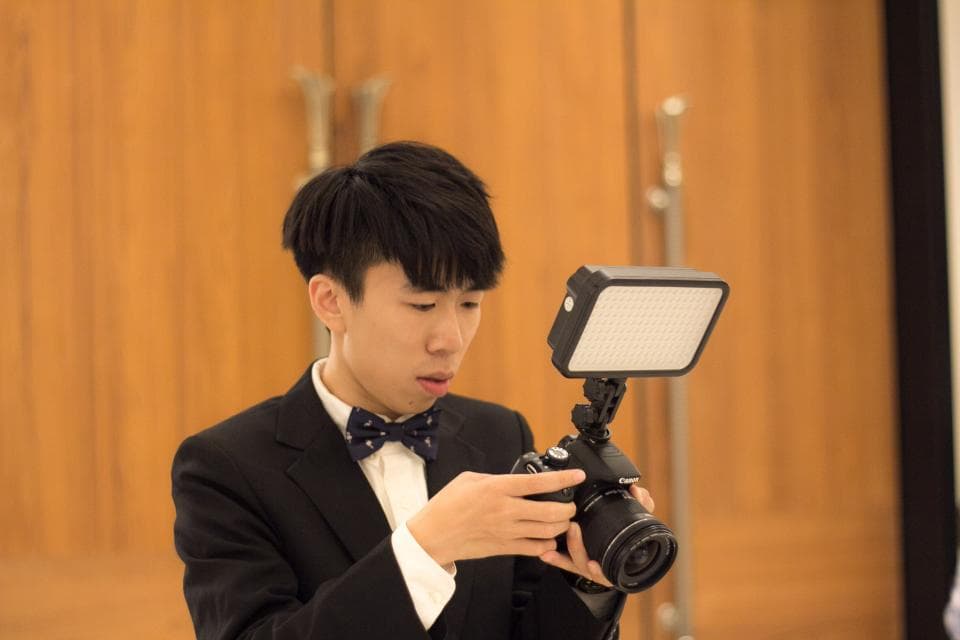 1) 照片中是一位年輕男子,他穿著西裝,打著領結,正在使用相機拍照,背景是一面木板牆