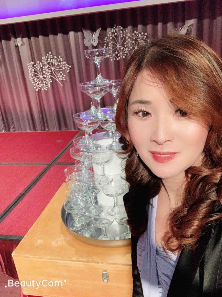 這張「女人站在香檳塔旁邊」的照片是,一個穿著深色外套的女人站在一個用很多香檳杯組成的塔旁邊,她的頭髮是棕色,臉上帶著微笑。