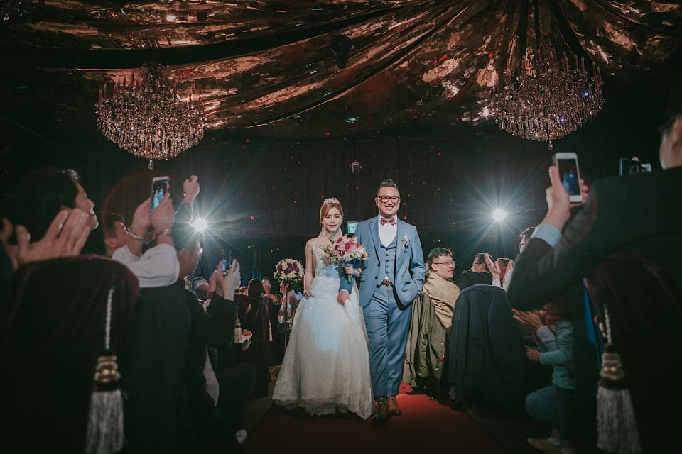 這張照片是一對新人正在步入禮堂,新郎穿著黑色西裝,新娘穿著白色婚紗,他們手牽著手,笑容滿面,看起來非常幸福。