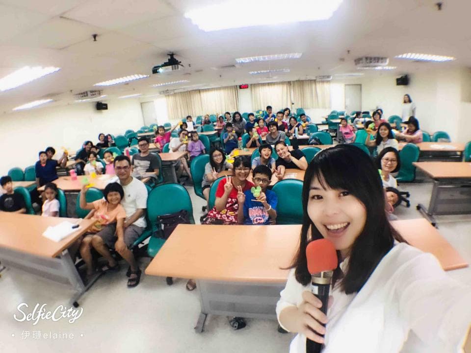 這張「一群人聚會」的照片是,一群人在教室裡聚會,其中一位女生拿著麥克風,看起來很開心的在講話,其他人在聽講。