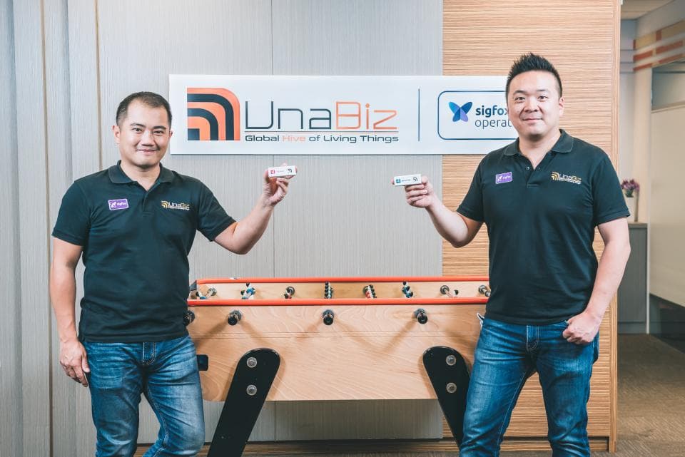 這張「UnaBiz」的照片是兩位男士站在足球桌前,他們都穿著黑色的T恤和牛仔褲,手裡拿著小型裝置。桌子後面的牆上有一個 UnaBiz 的標誌。