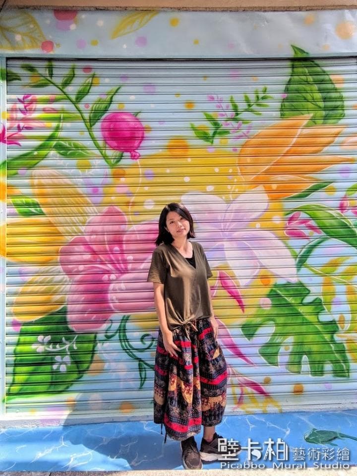 這張照片是一幅以花卉為主題的街頭壁畫,畫中有一位年輕女子站在壁畫前,她穿著一件簡單的T恤和一條色彩繽紛的長裙,襯托出壁畫的鮮豔色彩和豐富細節。