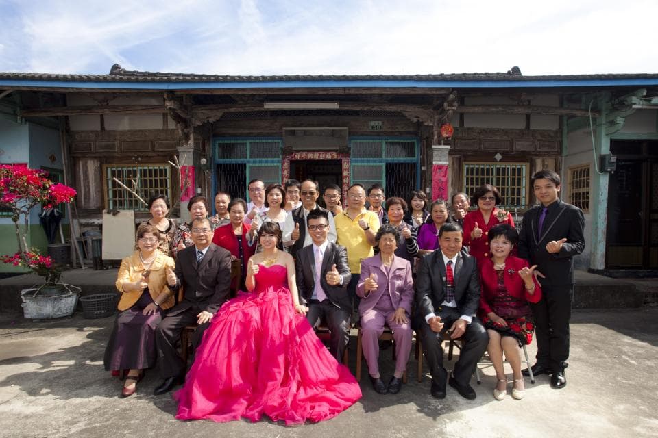 這張照片是一對新人結婚的照片,他們站在一棟老房子前面,周圍是他們的親朋好友。新郎穿著黑色西裝,新娘穿著粉紅色禮服,他們顯得非常幸福和喜悅。