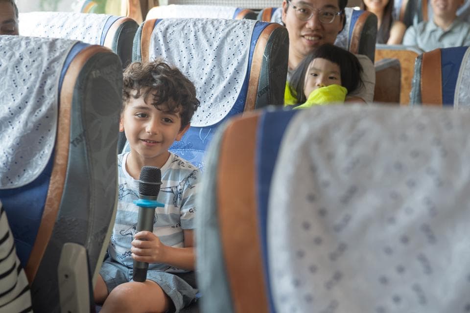 這張「公車」的照片是一個男孩坐在座位上,手拿著麥克風。他身後坐著一個大人,一個小女孩坐在前面,看起來她們都很開心。