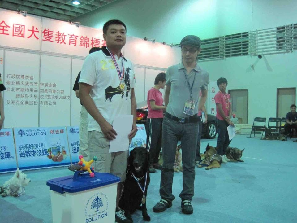 這張「訓練狗狗」的照片是,一位年輕男子正在訓練一隻黑色狗狗,他身穿藍色短袖上衣,身邊還有一位女士在旁邊觀看,背景是室內場地,以及一些其他訓練狗狗的場景。