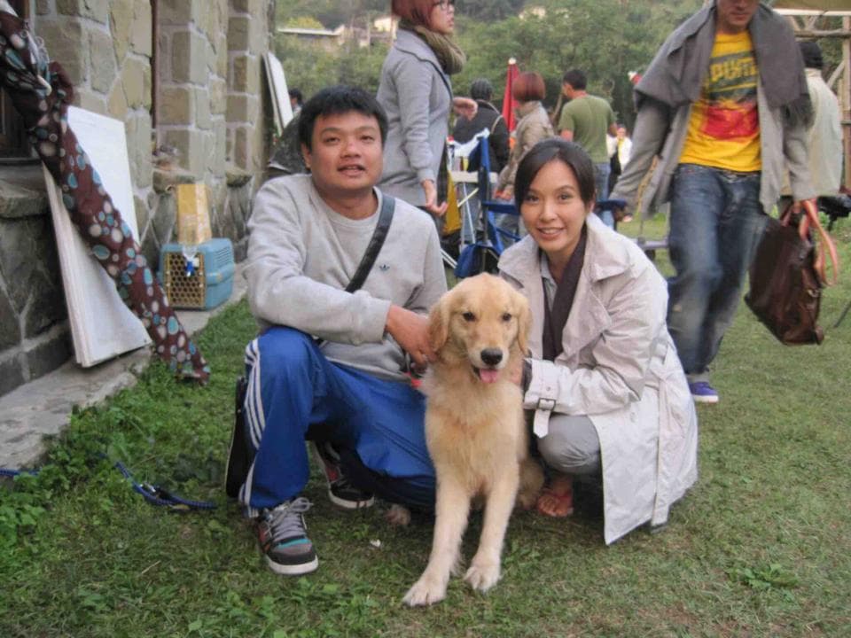 這張照片是一隻可愛的金毛獵犬,它正被一位男士和一位女士擁抱著。牠的尾巴搖擺,嘴巴微張,看起來非常開心。