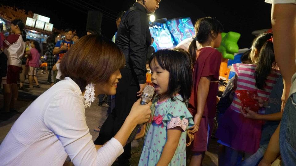 這張照片是一個年輕女性正在對一個小女孩說話。年輕女性手持麥克風,而小女孩則看著麥克風,似乎正在準備說話。