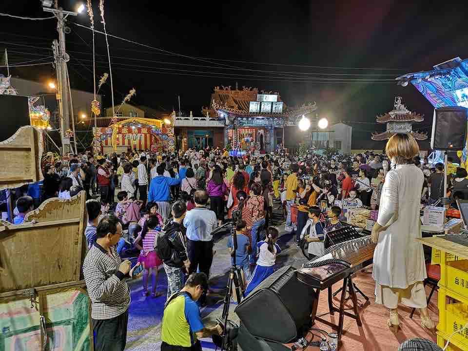 這張照片是一群人聚集在廟宇前,其中一個穿著白色衣服的女性在演奏樂器,吸引著人們的目光。