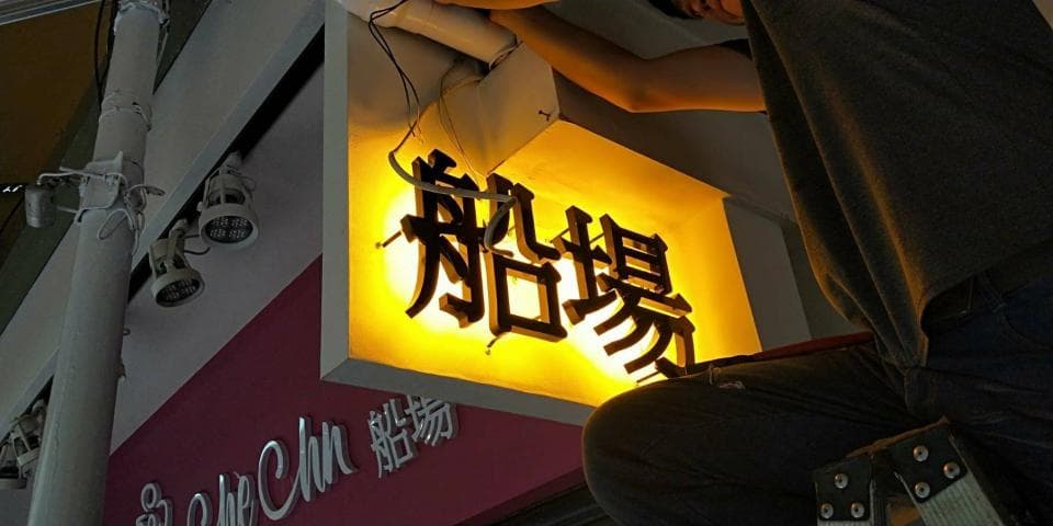 這張照片是一個正在安裝發光招牌的工人。招牌上用黑色字體寫著「奶茶」兩個字,招牌被黃色的燈光照亮,看起來非常醒目。