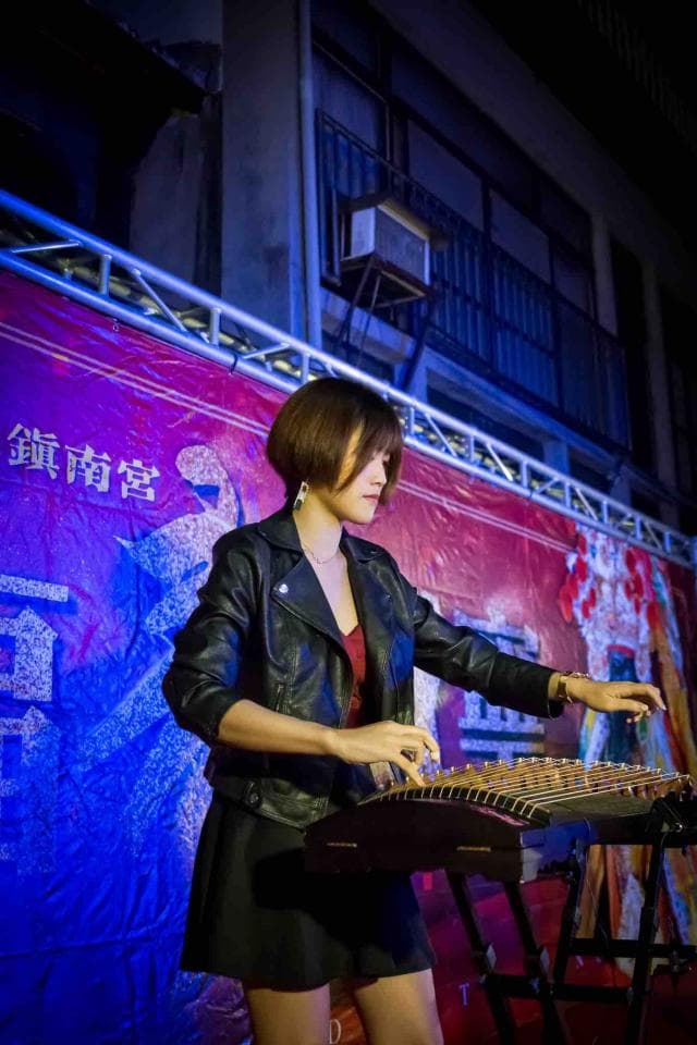 這張「一位年輕女子在舞臺上彈奏古箏」的照片是。她穿著黑色皮衣,裙子,正專注地彈奏著古箏。背景是一面紅色和藍色的布幕,上面寫著「金榜題名」。