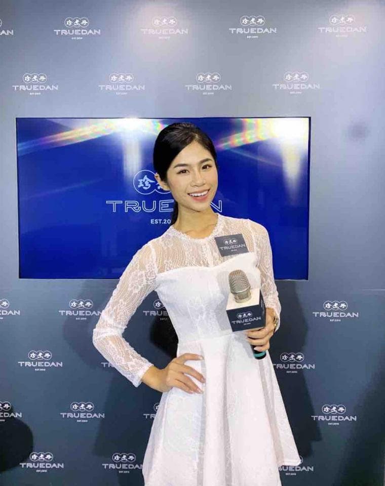 這張「美女手持麥克風站在TRUEDAN品牌牆前」的照片是,一個穿著白色蕾絲洋裝的年輕女性,站在一面印有「TRUEDAN」的品牌牆前,手持麥克風,露出自信的笑容。