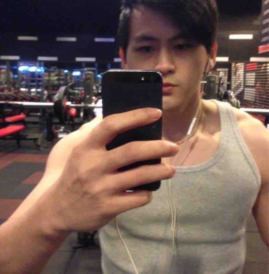 這張照片是一個年輕男子在健身房自拍。他穿著一件灰色的背心,戴著耳機,手裡拿著一部智慧型手機。他看起來很健壯,並且似乎很享受健身房的環境。